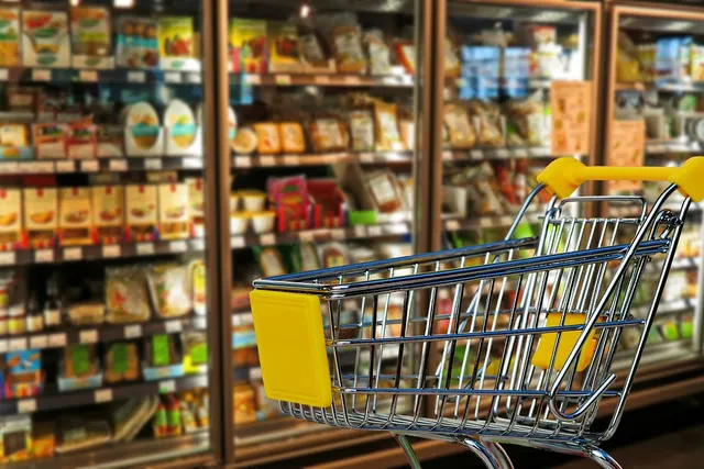 Empty shopping cart with yellow handle in a grocery store aisle beside refrigerated cases of packaged food