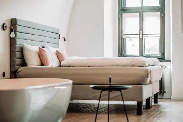 Bright bedroom with a bed with sage upholstered headboard, pink pillows, a small black side table and a white bathtub