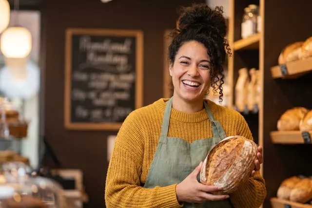 Smiling baker in a mustard sweater and green apron holds a sourdough loaf beside shelves of fresh bread