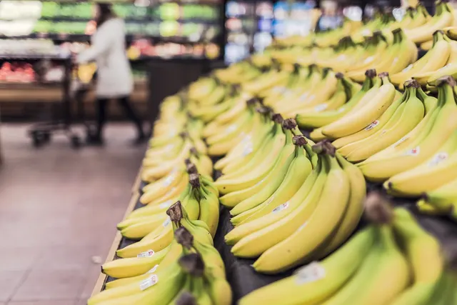 Rows of ripe bananas on a grocery store produce display, with a blurred shopper pushing a cart in the background