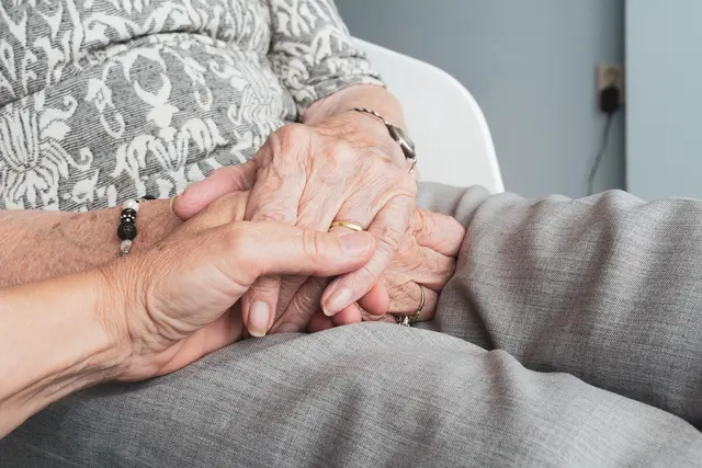 Caregiver holding the wrinkled hands of an elderly woman in a patterned blouse resting on her lap