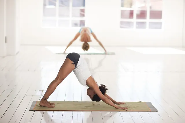 Two women practicing downward dog yoga poses on mats in a bright, white-floored studio with large windows