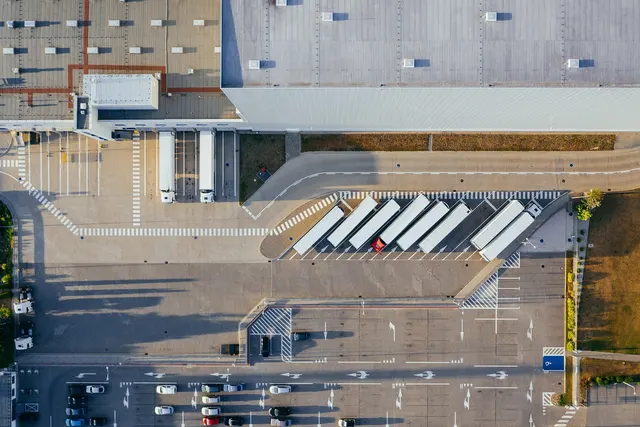 Aerial view of a warehouse with white semi-trailers parked at loading docks beside a car parking lot