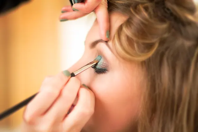 Makeup artist with green nail polish applying teal eyeshadow with a fine brush to a blonde woman's eyelid