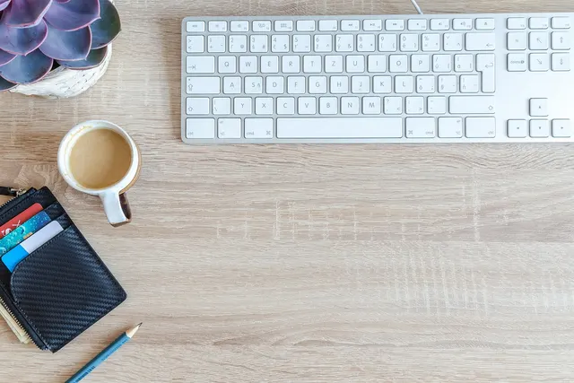Wooden desk with a white keyboard, cup of coffee, black wallet holding credit cards, pencil and a potted succulent