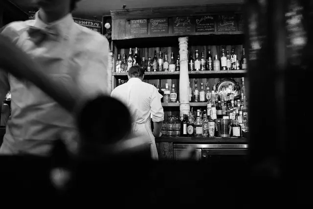 Black-and-white bar scene with a bartender in a white shirt and apron facing liquor shelves, a blurred server in front
