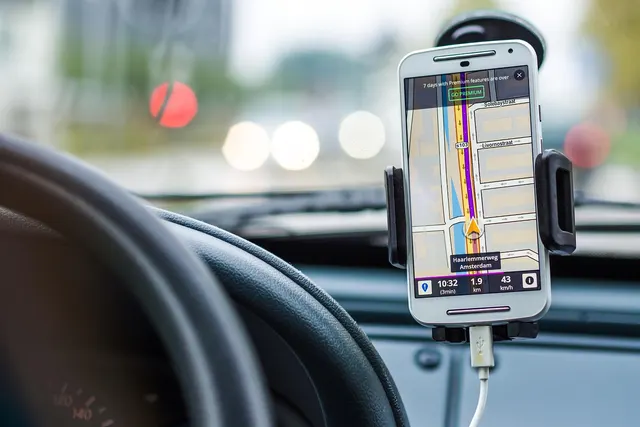 Smartphone in a dashboard mount showing GPS navigation, with a steering wheel and blurred traffic lights ahead