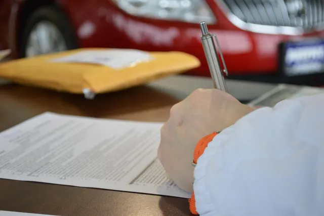 Customer signing a printed sales contract with a silver pen at a dealership desk, red car in the background