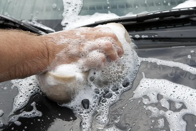 Close-up of a hand scrubbing a dark car hood with a soapy sponge near the windshield wipers