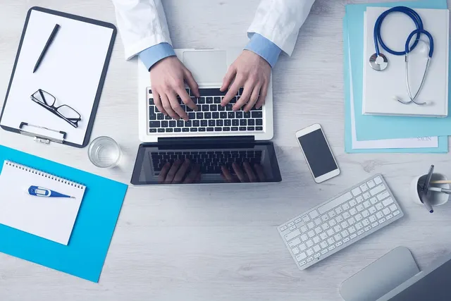 Overhead view of a doctor in a white coat typing on a laptop beside a stethoscope, smartphone, clipboard and thermometer