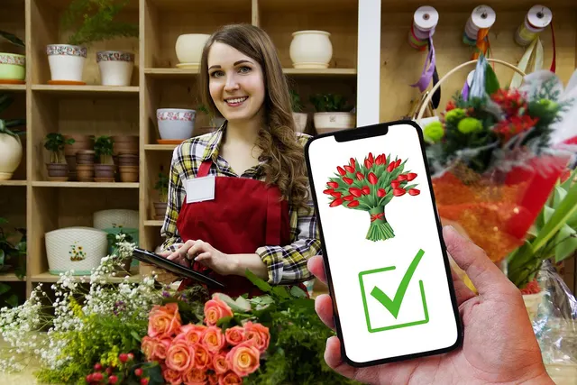 Smiling florist in red apron uses a tablet in her shop as a customer's phone shows a confirmed online bouquet order