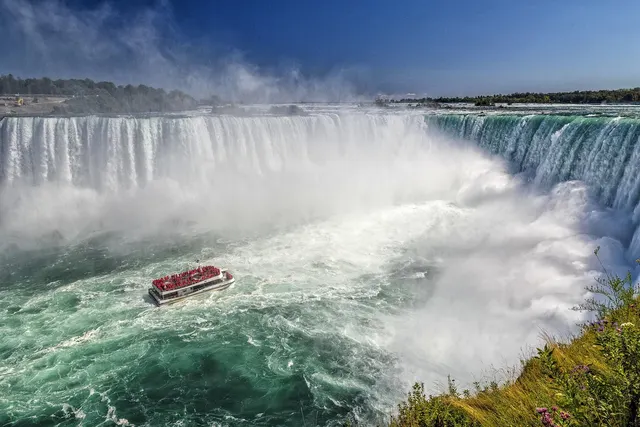 Tour boat full of passengers in red ponchos sailing through mist at the base of a huge horseshoe-shaped waterfall