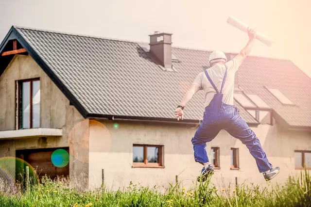 Contractor in blue overalls and hard hat leaps in celebration, holding rolled blueprints, outside a house under construction
