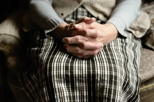 Close-up of an elderly woman's wrinkled hands clasped on her lap over a plaid skirt, wearing a gray cardigan