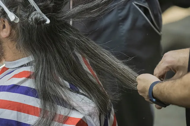 Stylist trimming a client's long gray-streaked hair, sectioned with clips, client wearing a striped salon cape
