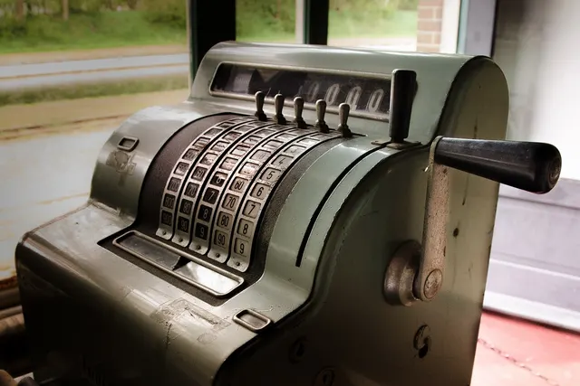 Vintage metal mechanical cash register with numbered keys and side crank handle, set beside a window