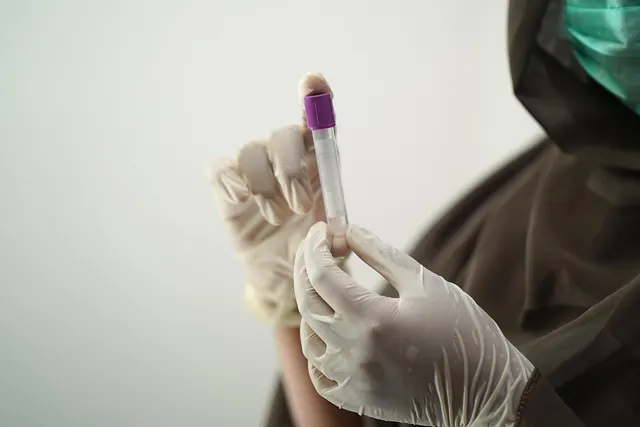Gloved hands holding a purple-capped test tube, a person in a green face mask and dark hood behind