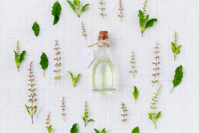 Corked glass bottle of clear oil with a blank tag, surrounded by fresh basil leaves and flower sprigs on white linen