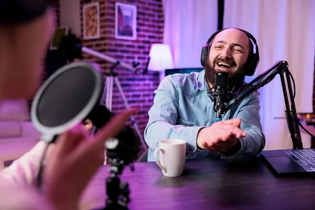 Bearded man in headphones laughs into a podcast mic in a neon-lit home studio, with a guest's pop filter blurred in front