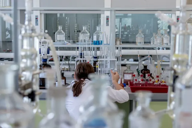 Lab technician in a white coat working at a bench lined with glass bottles, flasks and sample vials in a laboratory