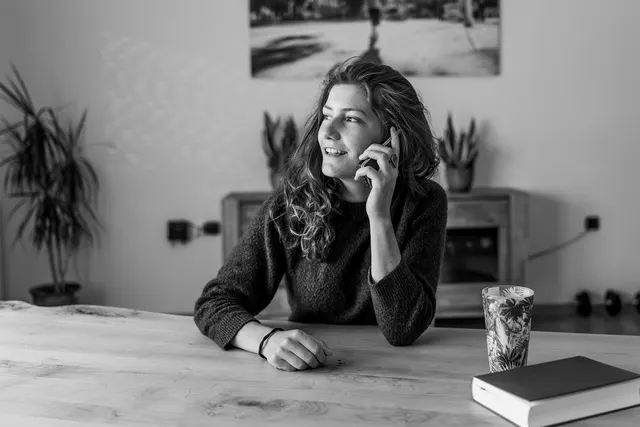 Smiling woman with curly hair talking on her phone at a wooden table, with a book and cup nearby, in black and white