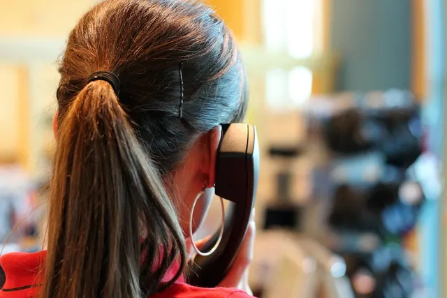 Woman with a long ponytail and hoop earring, seen from behind, talking on a desk phone handset in a blurred retail shop