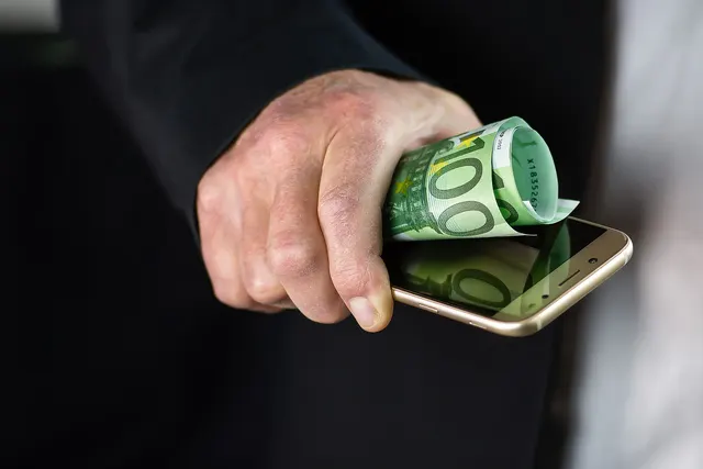 Close-up of a hand in a dark suit sleeve holding a rolled 100 euro banknote and a gold smartphone