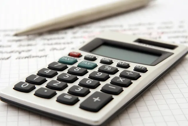 Silver desktop calculator with TAX+ and TAX- keys on grid paper beside a pen and blurred handwritten notes