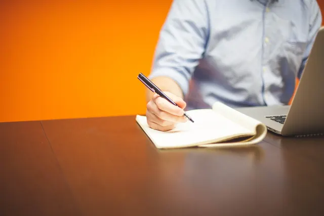 Person in a light blue shirt writing notes on a notepad beside a laptop at a wooden desk against an orange wall