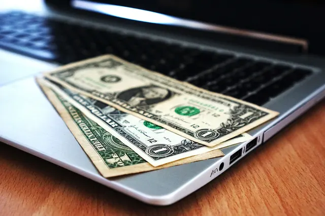 Three US one-dollar bills resting on a silver laptop keyboard on a wooden desk