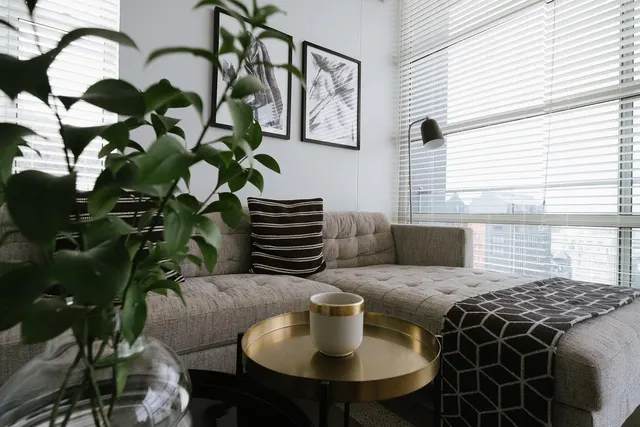 Modern guest suite living area with gray tufted sectional sofa, gold tray side table with cup, potted plant and window blinds