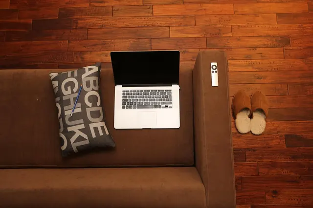 Overhead view of a brown sofa with an open laptop, alphabet pillow and remote, slippers on a wood floor