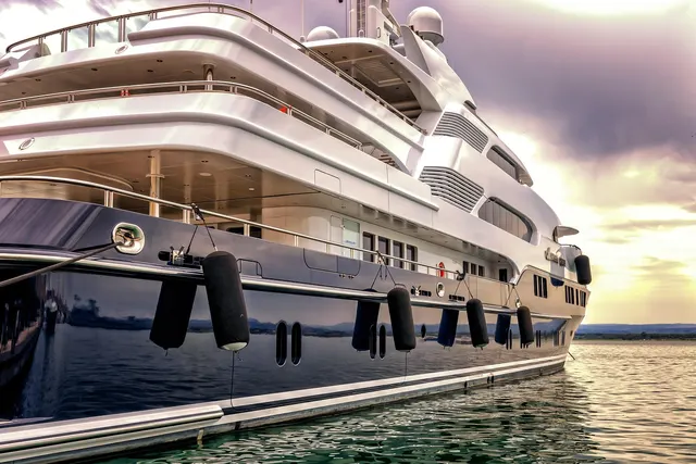 Large white luxury motor yacht moored in calm water with black fenders along its dark hull under a sunset sky