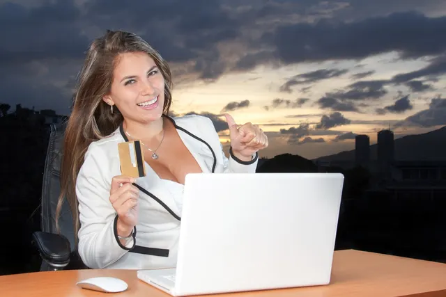 Smiling woman in a white blazer holds a gold payment card and gives a thumbs up behind a white laptop at dusk
