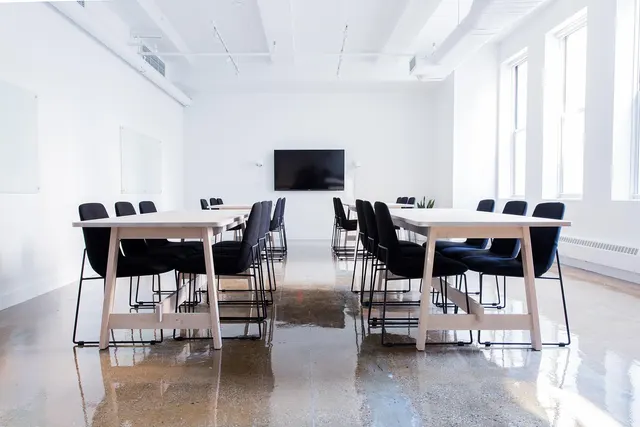 Empty bright meeting room with two light wood tables, black chairs, a wall-mounted TV and polished concrete floor