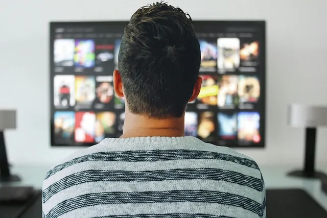 Man in a striped sweater, seen from behind, browsing a grid of movie thumbnails on a streaming service on a TV screen