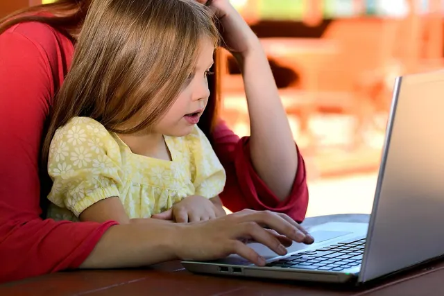 Mother in a red top types on a silver laptop at a wooden table while her young daughter sits on her lap watching