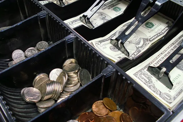 Open cash register drawer holding US dollar bills under metal clips and compartments of quarters, nickels and pennies