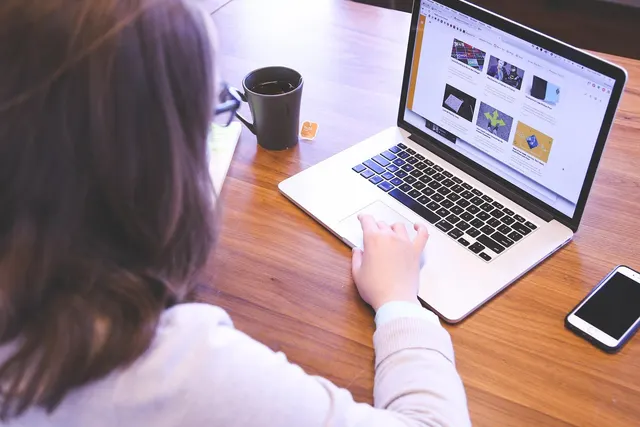 Over-the-shoulder view of a woman browsing a website on a laptop at a wooden desk with a coffee mug and smartphone