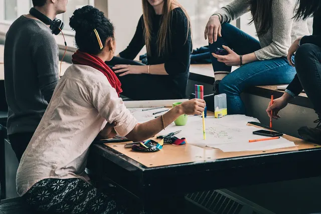Young adults gather around a worktable with large paper sheets, colored markers and phones during a group study session