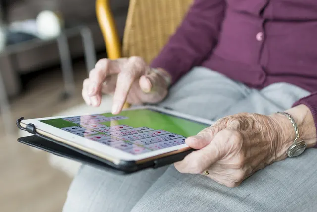 Close-up of an elderly woman's hands holding a tablet showing a solitaire game while seated in a wicker chair at home