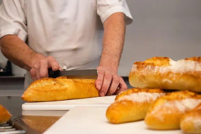 Baker in a white chef's coat slicing a fresh baguette on a white cutting board, with crusty loaves in the foreground