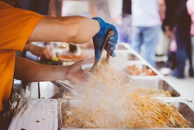 Food vendor in blue gloves using tongs to serve steaming noodles from trays into a takeout container at an outdoor event