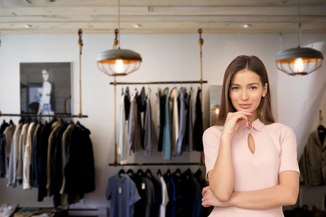 Woman in a pink dress posing thoughtfully in a clothing boutique with garments on hanging rails and pendant lamps