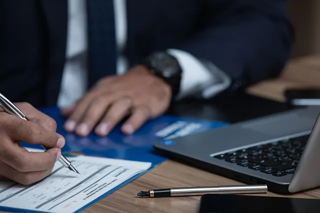 Person in a dark suit signing a paper form with a silver pen at a wooden desk beside a laptop and blue folder