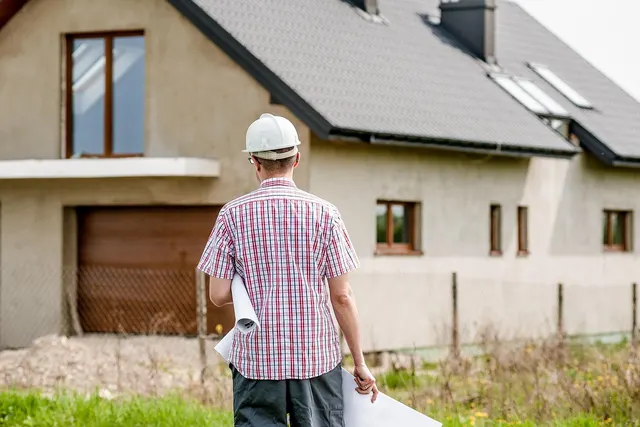 Contractor in white hard hat and plaid shirt holding rolled blueprints, facing an unfinished two-story house