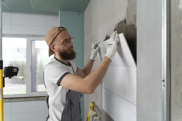 Bearded contractor in cap and gloves setting large white tiles on an adhesive-coated wall during a bathroom remodel