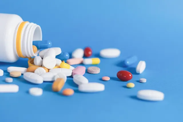 White prescription bottle tipped over with assorted white, blue, red and yellow pills and capsules spilling onto a blue surface