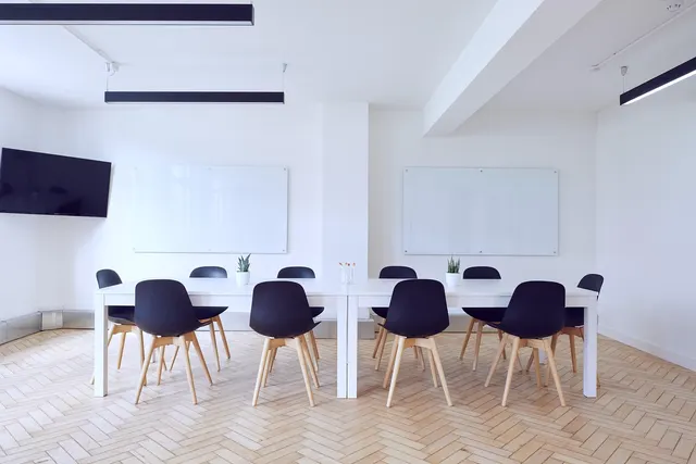 Empty bright meeting room with white tables, black chairs, wall-mounted TV and whiteboards on herringbone floor