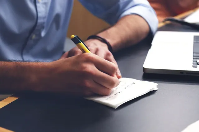 Man in a blue shirt writing notes with a yellow pen on a small paper beside a laptop on a dark desk